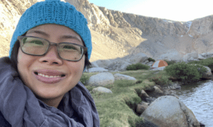 A close up picture of Tuyen out enjoying nature. She is smiling while wearing a blue knitted hat, glasses, and a gray scarf.  